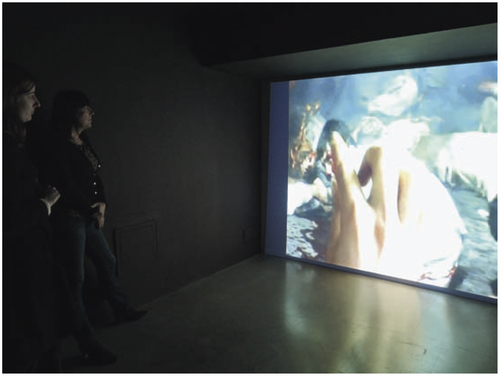 Viewers standing in a dark space, in front of a projected image filling the wall, showing a large hand pointing to a photograph behind.