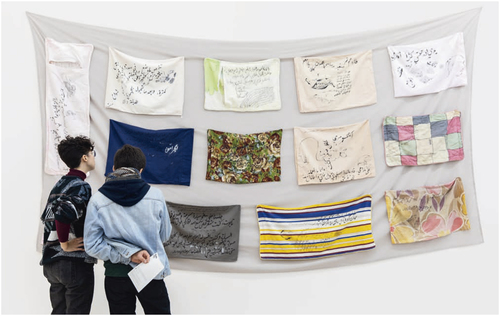 Viewers looking at a hanging grey bedsheet with several multicolored pillowcases stitched on top, some of which feature drawings and cursive Arabic inscriptions.