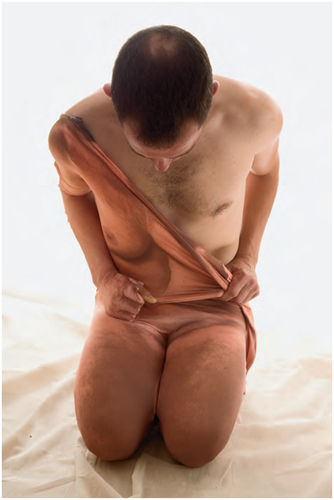 A naked male kneeling and bowing head while struggling to remove a full body suit. Half of the torso and upper body are exposed while half is still covered with the suit. The torso appears to be female. It is thus a man in a woman's skin.