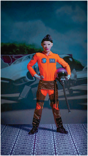 A figure in an orange pilot's suit, wearing a white mask, black boots on a patterned floor holding a helmet in right hand. In the background, photographed scenery of a military airforce base.