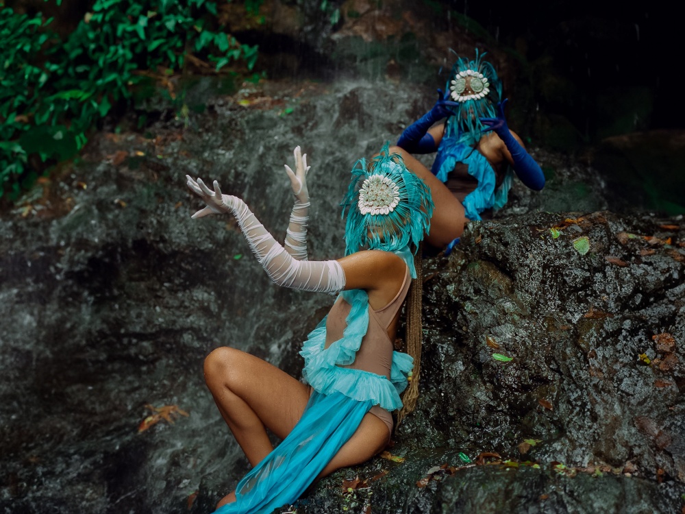 Two figures with their faces covered by masks, dressed in bodysuits adorned with shades of blue, sporting opera-length gloves.