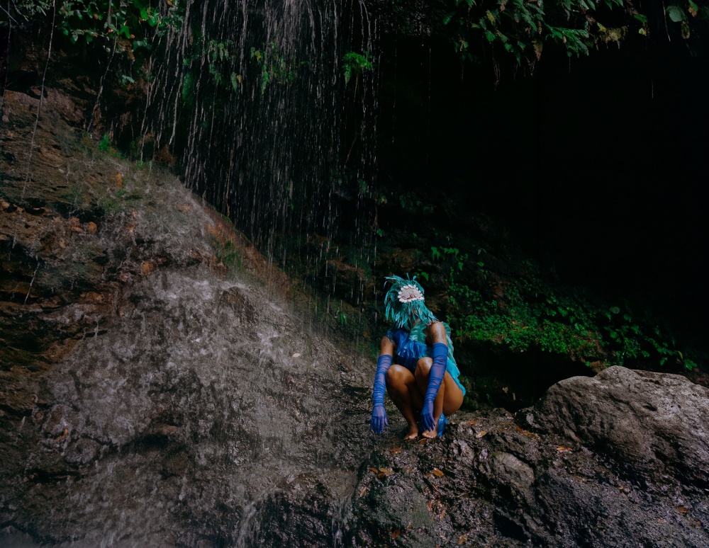A person crouches on top of the rocks of a waterfall, wearing a mask, blue opera-length gloves, and a bodysuit.