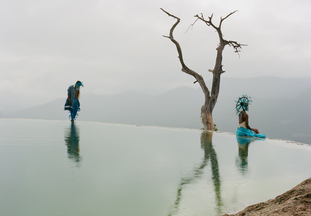 Two people with a tree in between them and a hot spring in the foreground.