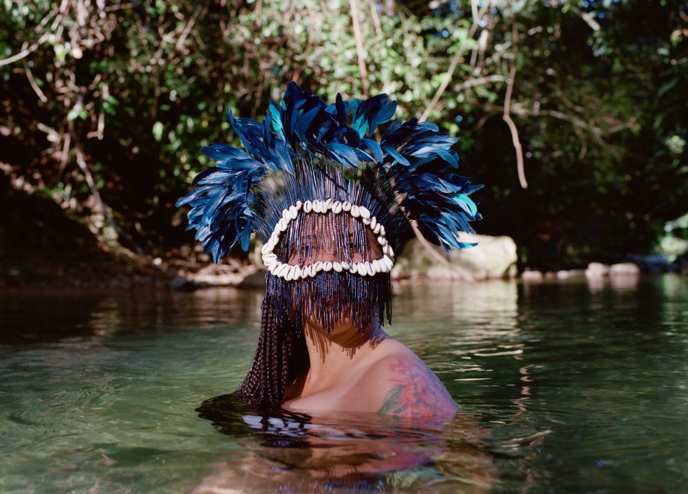 Portrait of a person wearing a mask adorned with blue feathers and cowrie shells inside a river from the shoulders down.