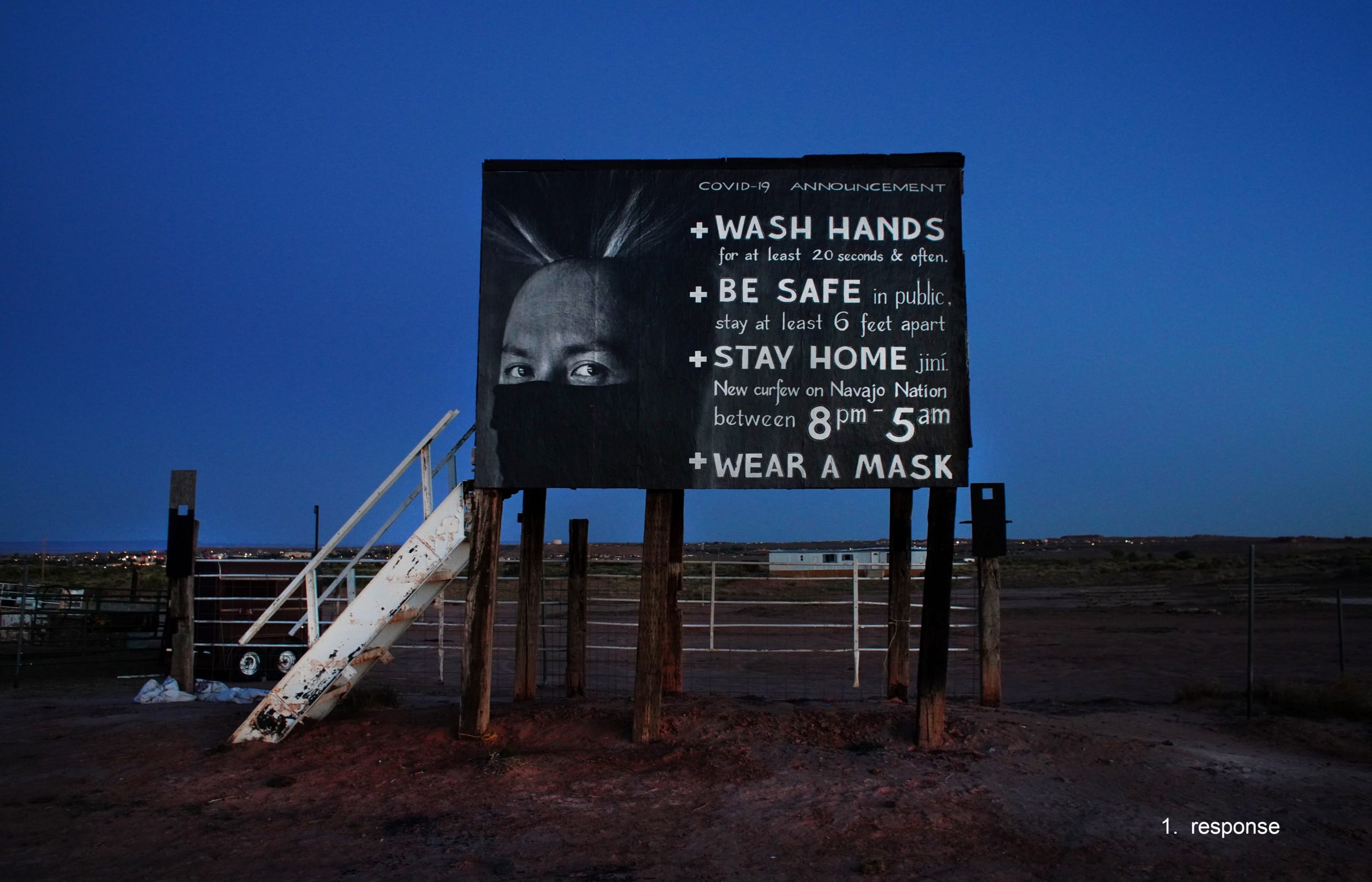 Color photograph, taken in pre-dawn light, of the Diné COVID PSA Poster (featuring the face of a Navajo man and instructions for staying safe from the coronavirus), installed on of a rodeo announcer's stand on the Navajo nation in Tuba City