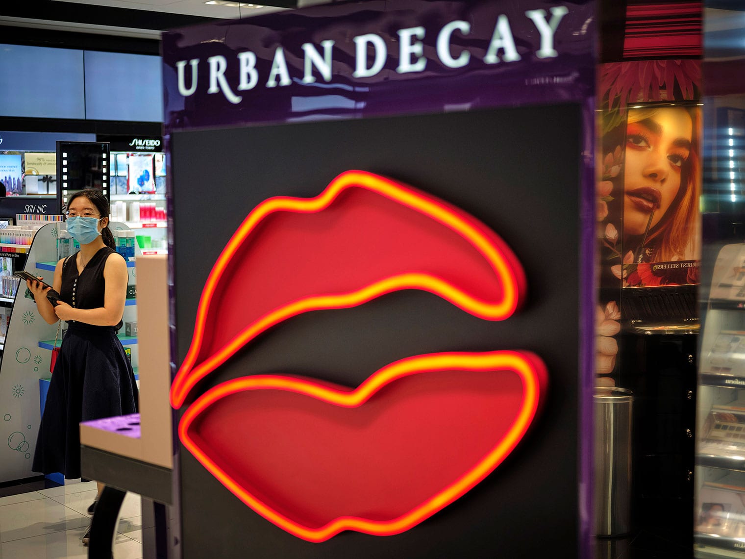 Sign with neon-outlined parted lips that reads "Urban Decay" at entrance to Sephora store, with masked woman in background looking at the camera