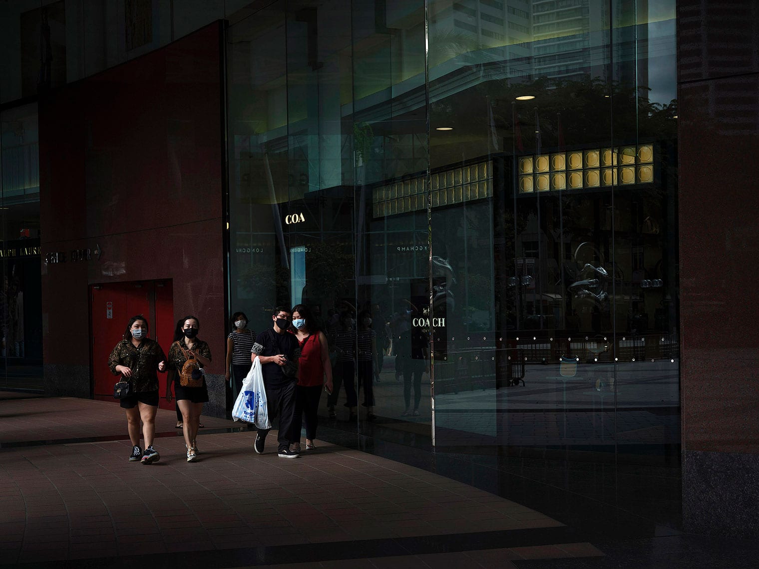 Masked shoppers walk along a darkened street beside a glass "Coach" shop window