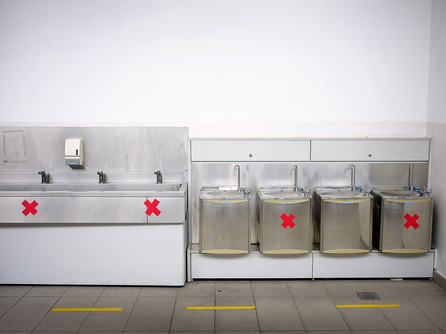 Seven water fountains lined up against the wall; every other foundatin is marked with a red X in tape
