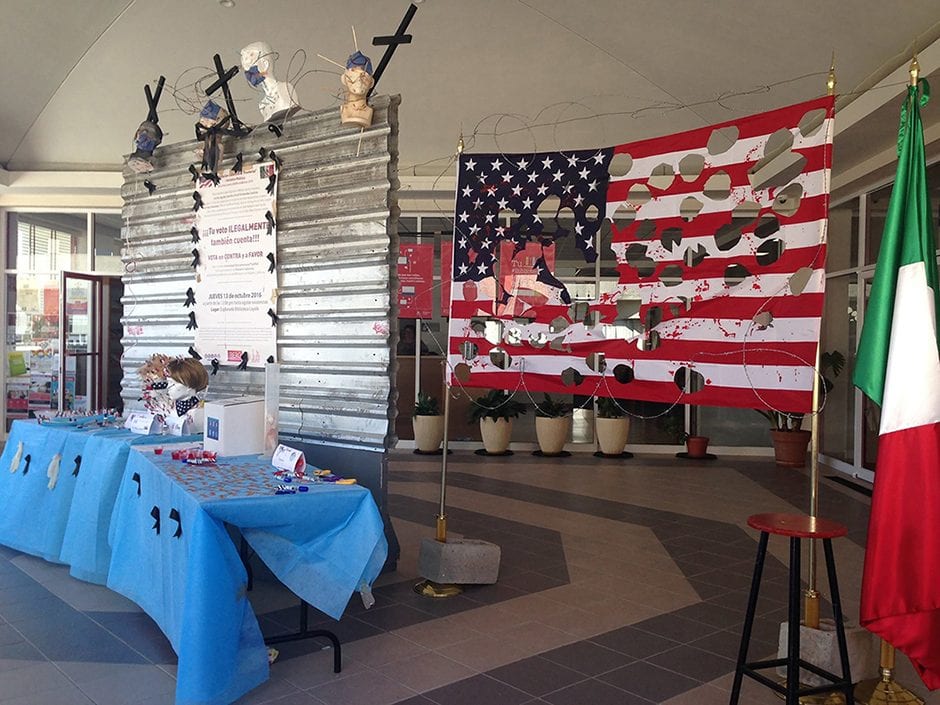 A table with ballots and a ballot box in front of a metal wall topped with crucifixes and barbed wire, next to an American flag with holes cut out of it