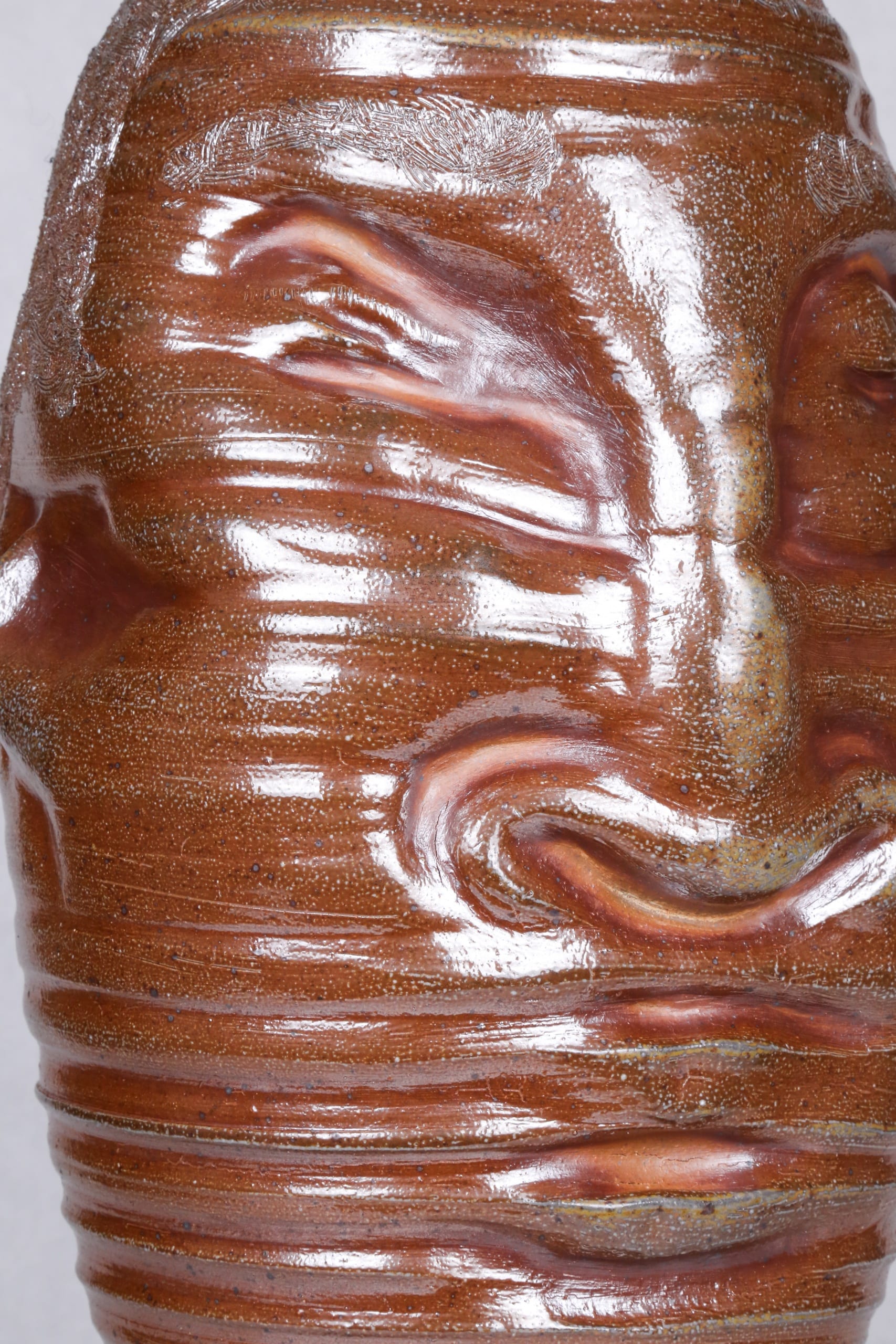 Color photograph of an abstract ceramic portrait head with distictive ridges wrapping around the circumfrance of the head, showing an up-close view of the object's front so the textures of the clay and the glaze are more visible. The object is monochromatic (warm brown) with a light, luminescent glaze applied to its surface. It appears as though it was made on a spinning potter‘s wheel.