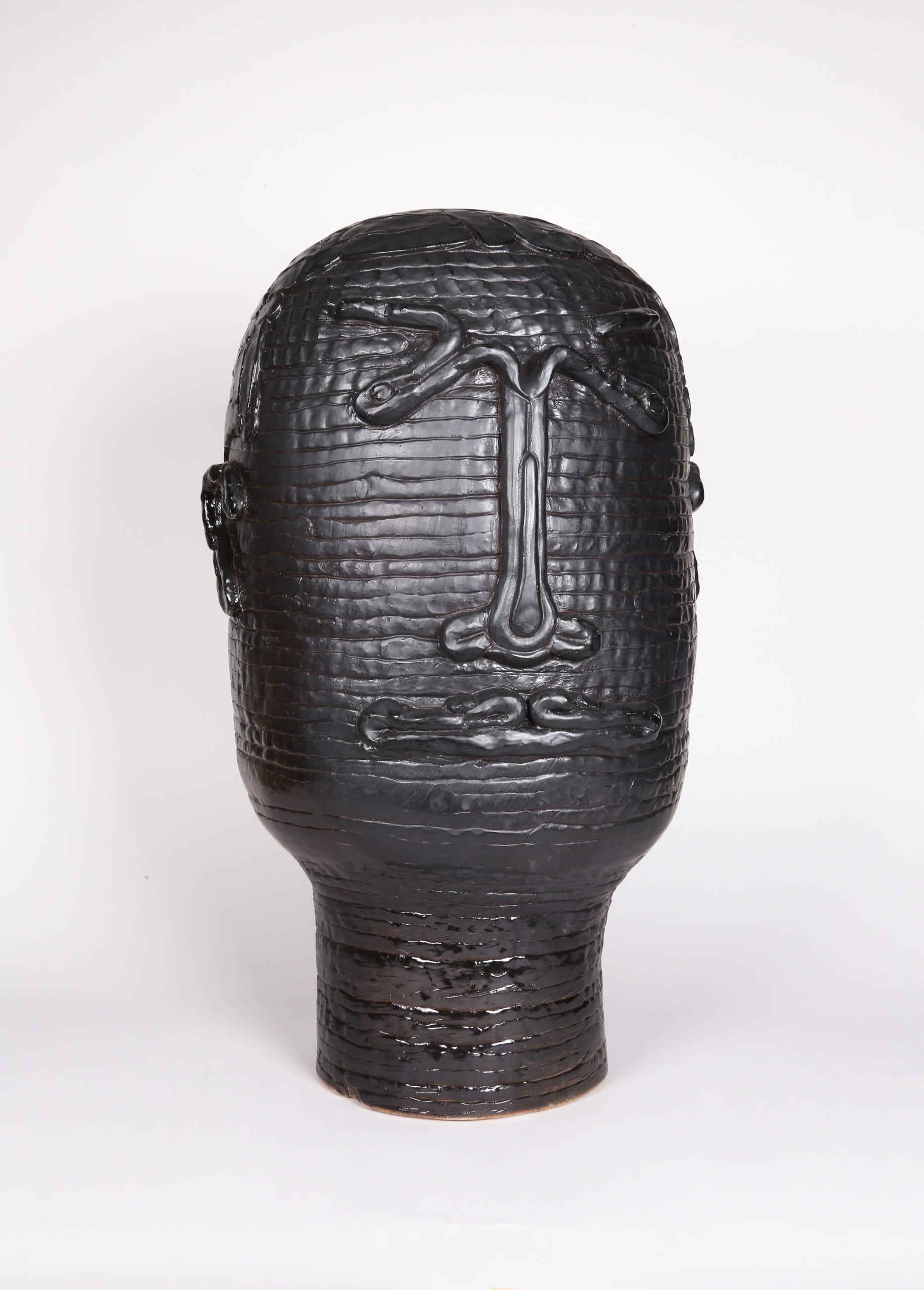 Color photograph with a frontal view of an abstract ceramic portrait head that appears to be made from a long coil of clay. The outline of two eyes, a nose, a mouth, and two ears are affixed to its surface. The object is a monochromatic black with a slight sheen to the glaze.