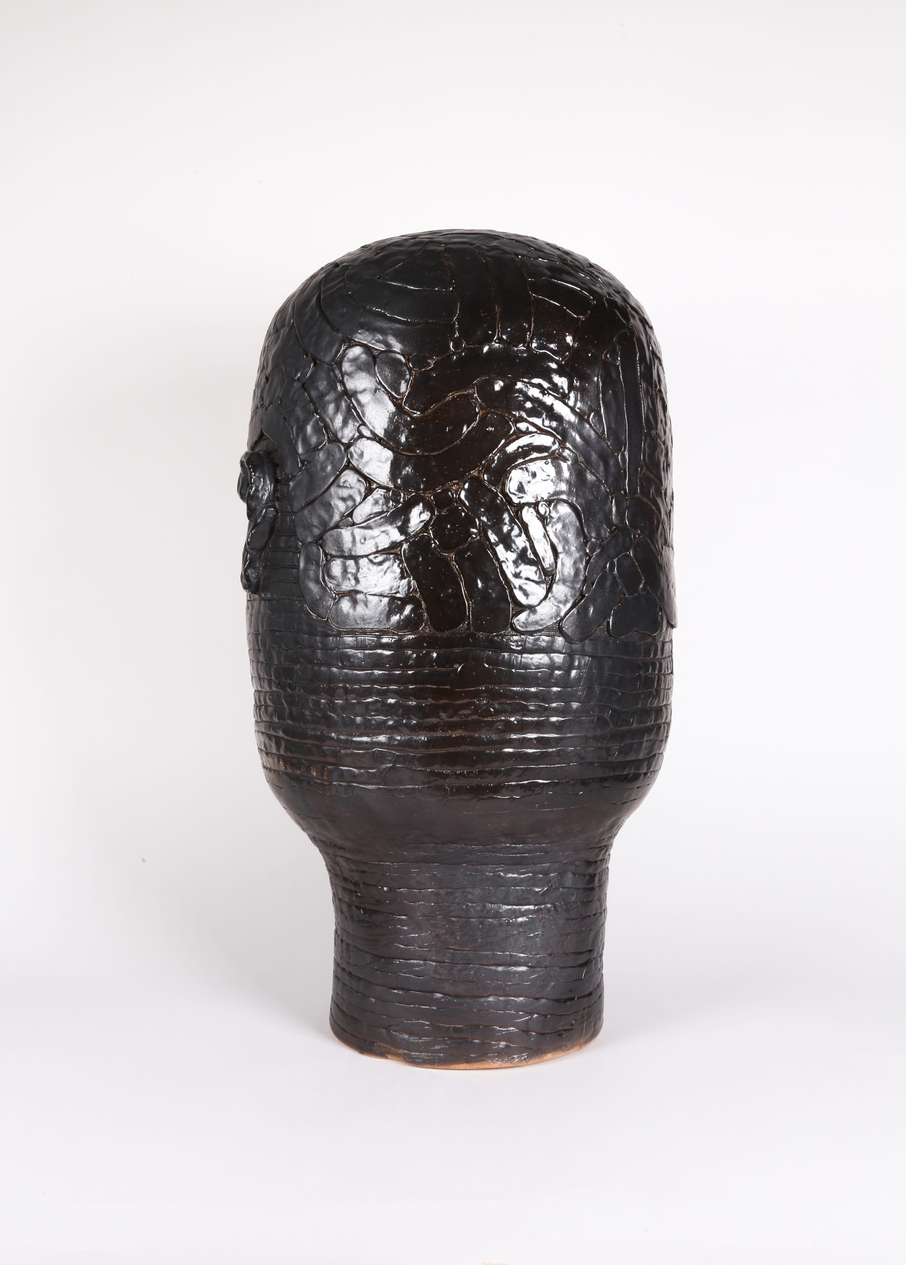Color photograph with a back view of an abstract ceramic portrait head that appears to be made from a long coil of clay. The outline of hair is affixed to its surface. The object is a monochromatic black with a slight sheen to the glaze.