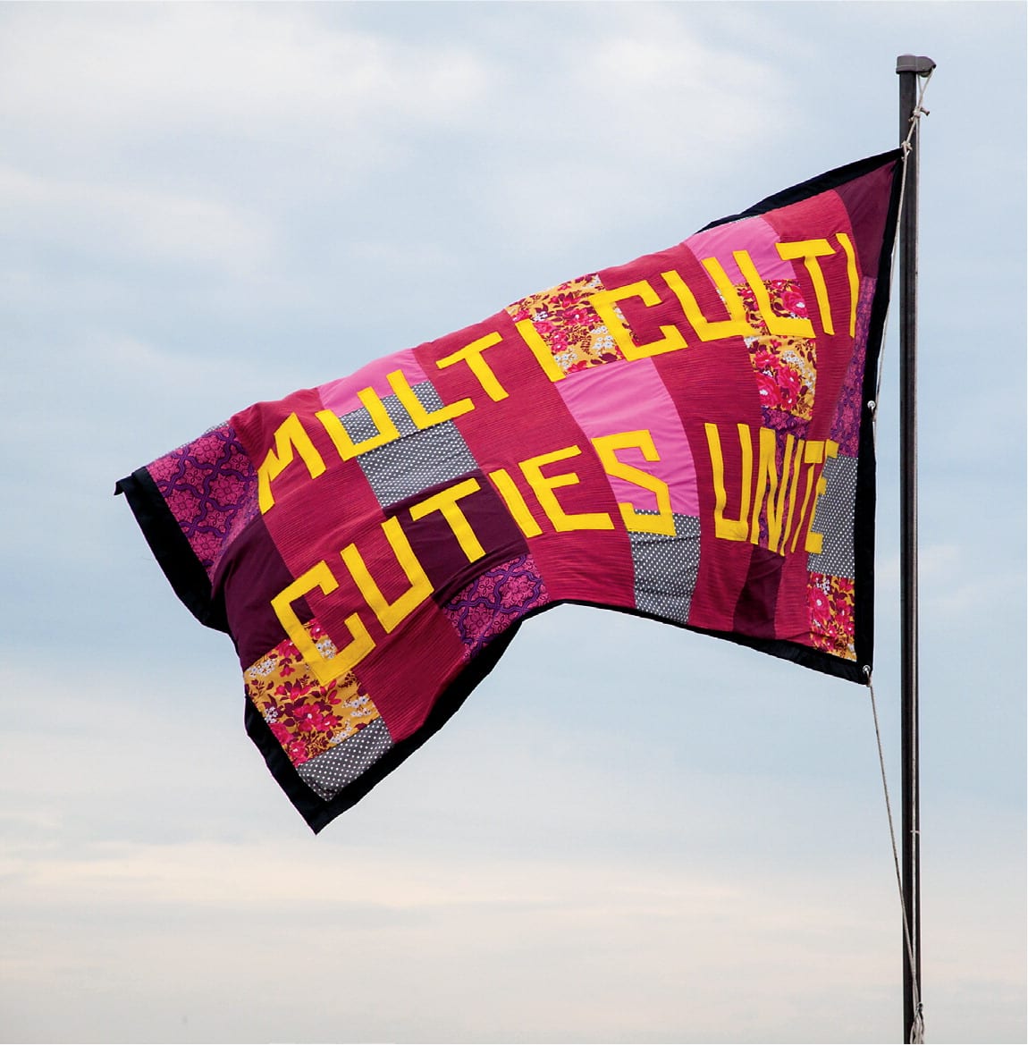 A flag made up of patchwork squares in various shades of hot pink, burgundy, and various patterns flies from a flagpole. Bold yellow text on the flag reads "Multi Culti Cuties Unite."