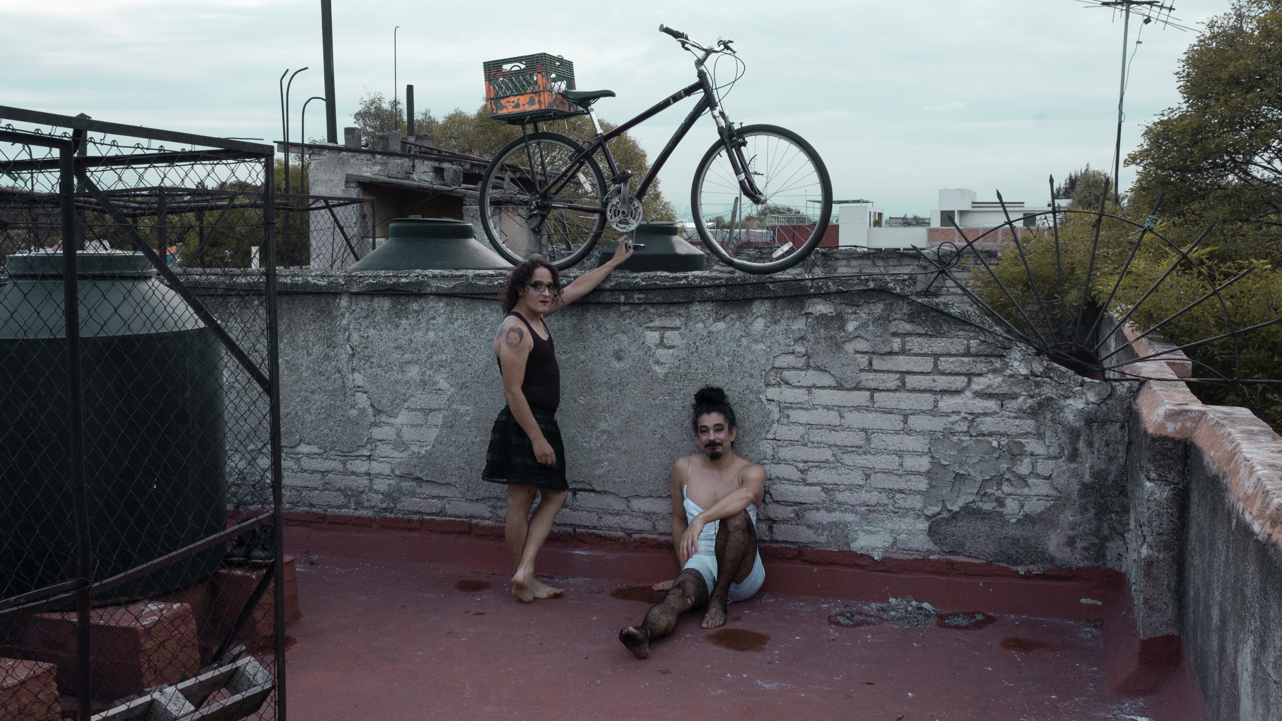 A gender-nonconforming person, standing, and a nonbinary person (the artist), sitting with one leg outstretched, are on a rooftop in Mexico City. A bike held up by the gender-nonconforming person balances on the wall of the rooftop.