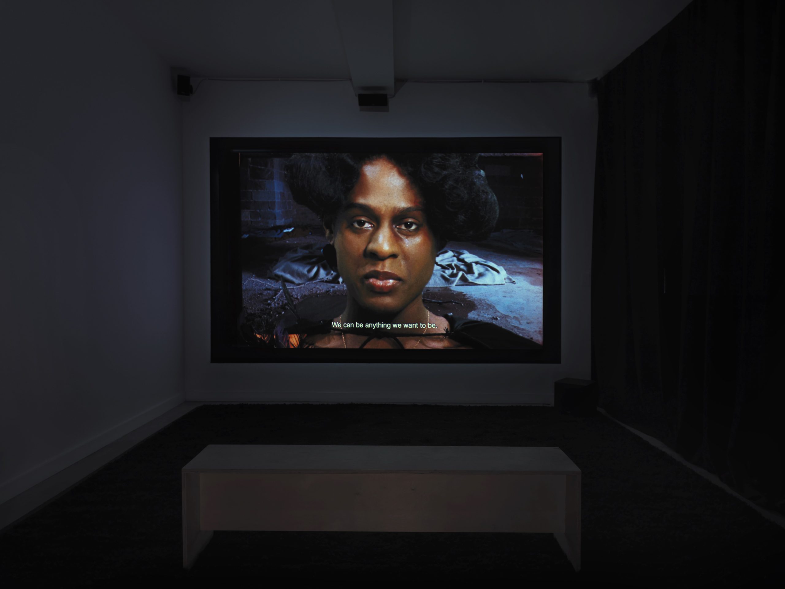 Photograph of a screening room with an image on screen of a close up face of a person with a tear falling from one eye, looking directly outward.